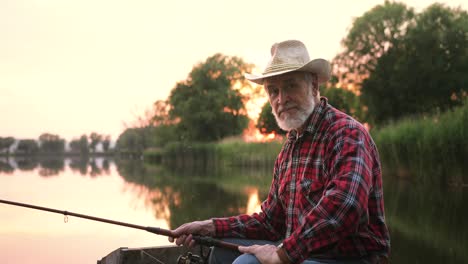 retrato de un pescador senior poniéndose sombrero y sentado en el muelle del lago mientras sostiene una caña de pescar al atardecer