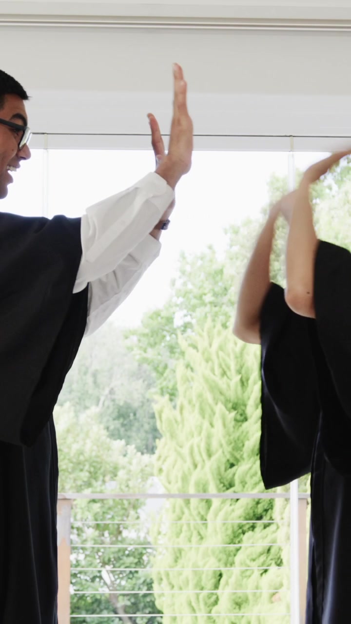 Diverse couple standing near big window putting black robes man in white shirt laughing, copy space