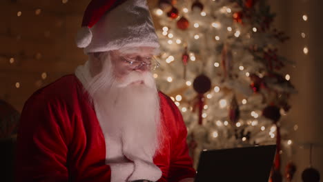 santa claus is working with a laptop at night in the light of christmas lights on the background of a christmas tree