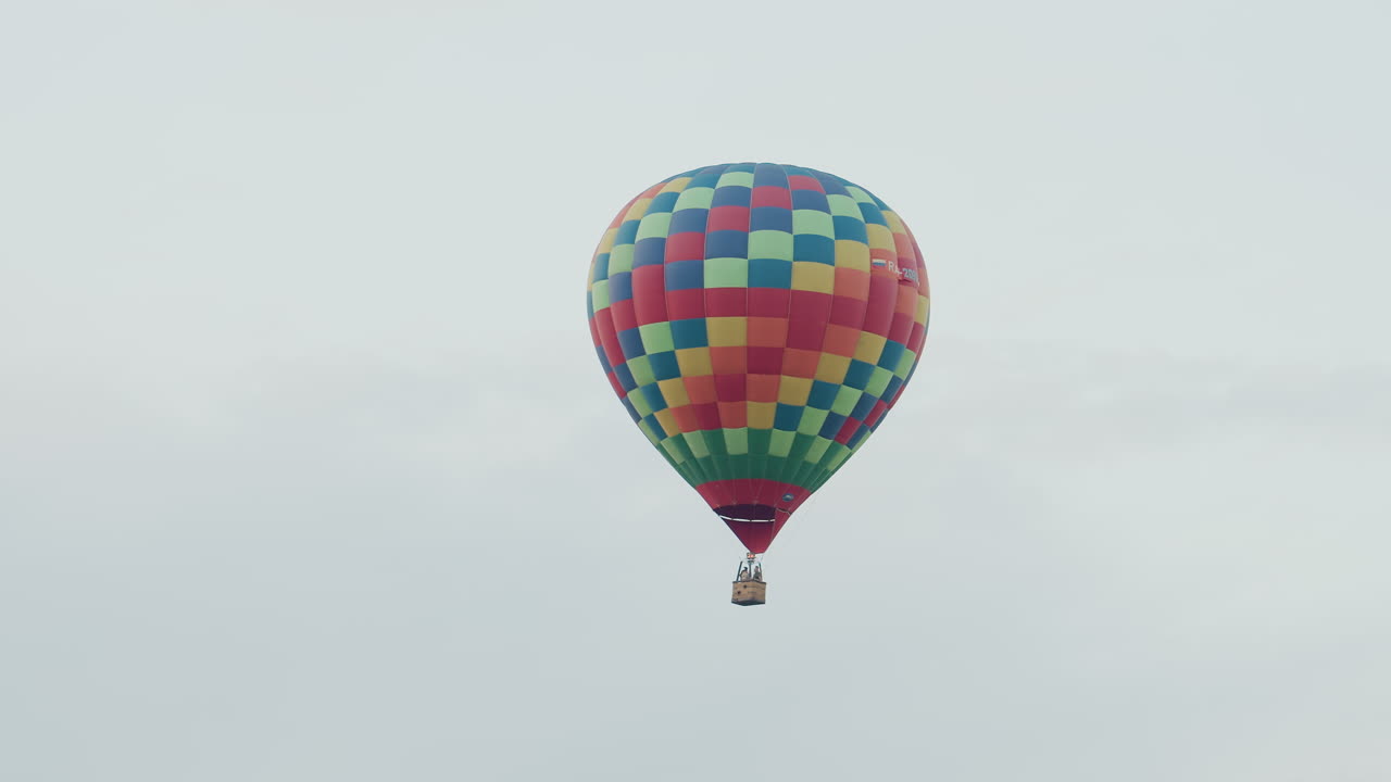 single passenger traveling in vibrant checkered hot air balloon drifting silently above green fields under soft pastel sky during morning flight adventure with distant balloon visible on horizon
