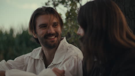 A man in period attire smiles while engaging in conversation outdoors with another person