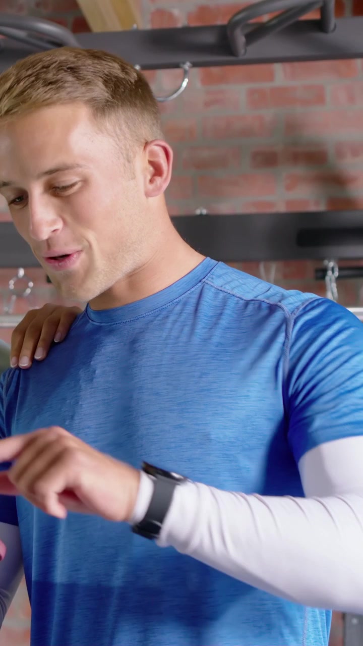 Vertical video: After partner tap, tapping man in blue tee checking plan on phone at gym with watch