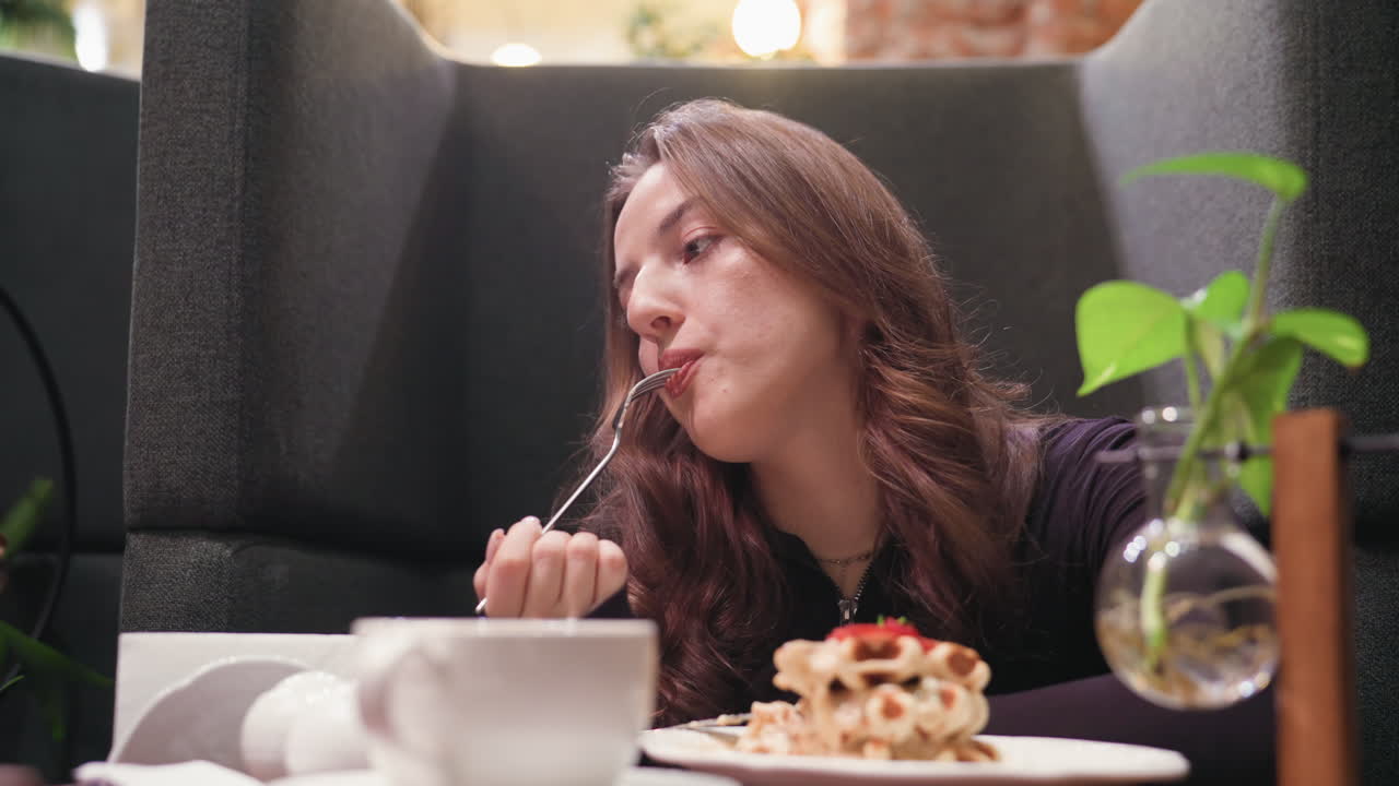 Lady gently eats waffles while appearing lost in thought, seated at small cozy table with warm decoration, soft lighting, white coffee cup, and table plant in calm relaxing environment
