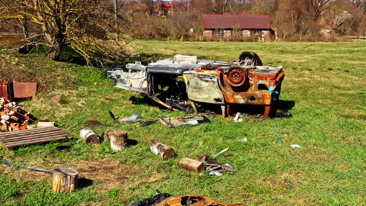Abandoned burned car lies overturned in grassy yard surrounded by scattered debris