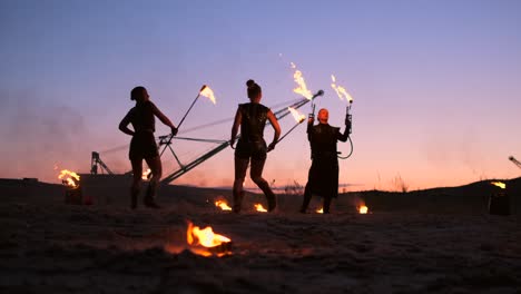 a group of men and woman fire show at night on the sand against the background of fire and tower cranes