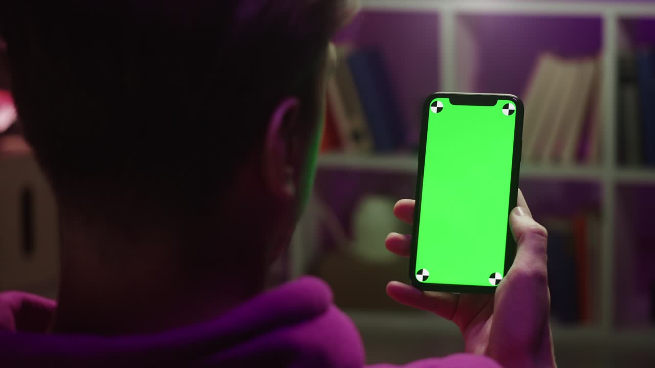 Man using smartphone with chroma key in evening. Young guy sitting on sofa and holding mobile phone with green screen, back view.
