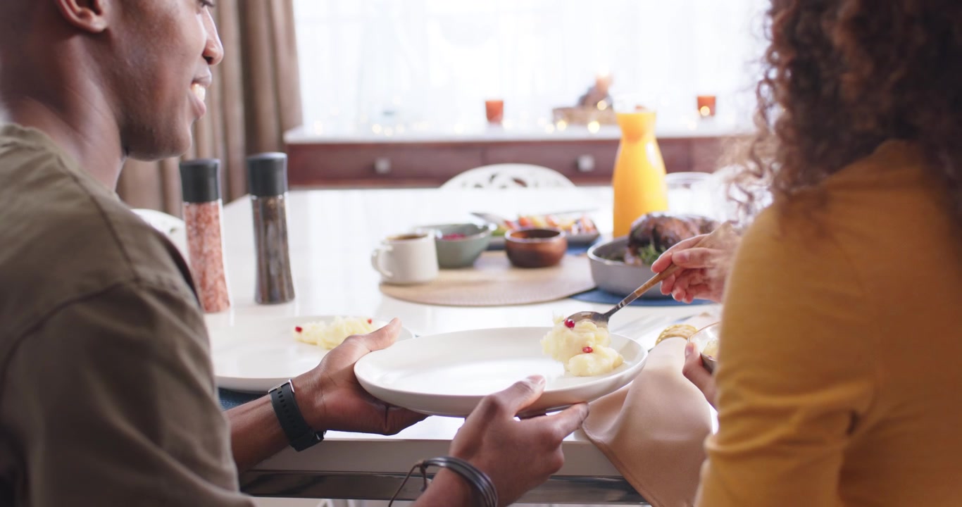Diverse couple serving meal at counter, man holding plate steady while woman spooning potatoes