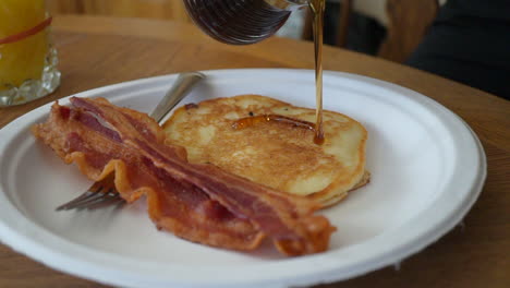 maple syrup being poured on a freshly fried pancake with two slices of bacon - slow motion