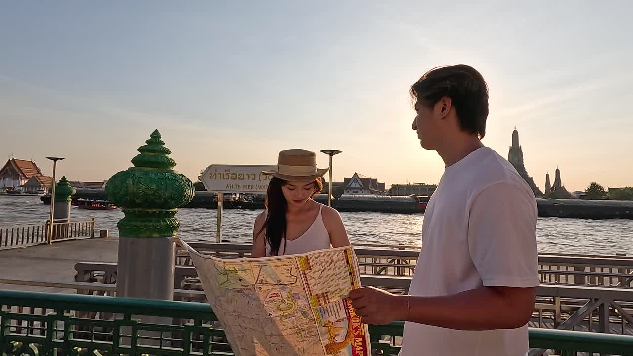 Cặp đôi trẻ du lịch xem bản đồ giấy tại bến tàu ven sông vào buổi hoàng hôn ấm áp, cảnh quay trung bình cầm tay.