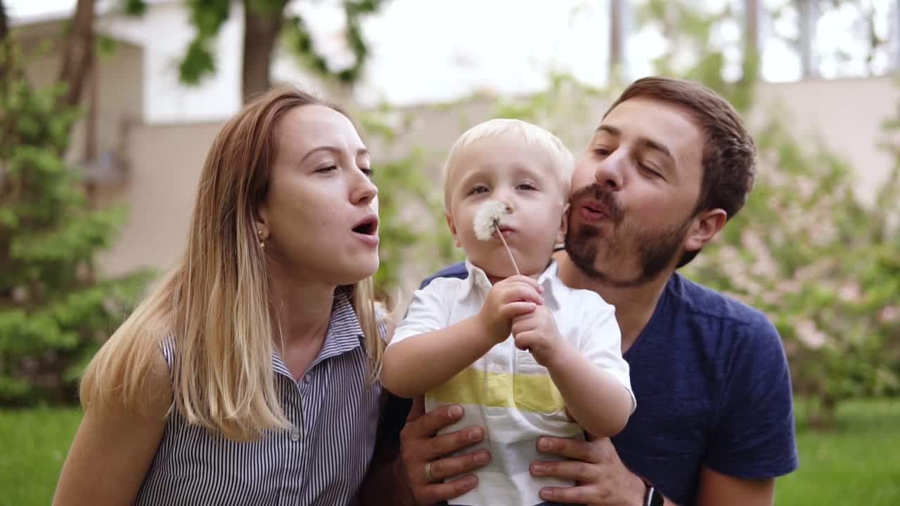 행복하고 즐거운 작은 아이가 중간에 있습니다. 부모님은 양쪽에서 소년에게 가습니다. 그들은 함께 꽃을 고 있습니다. 배경에는 초록색 잔디이 있습니다.