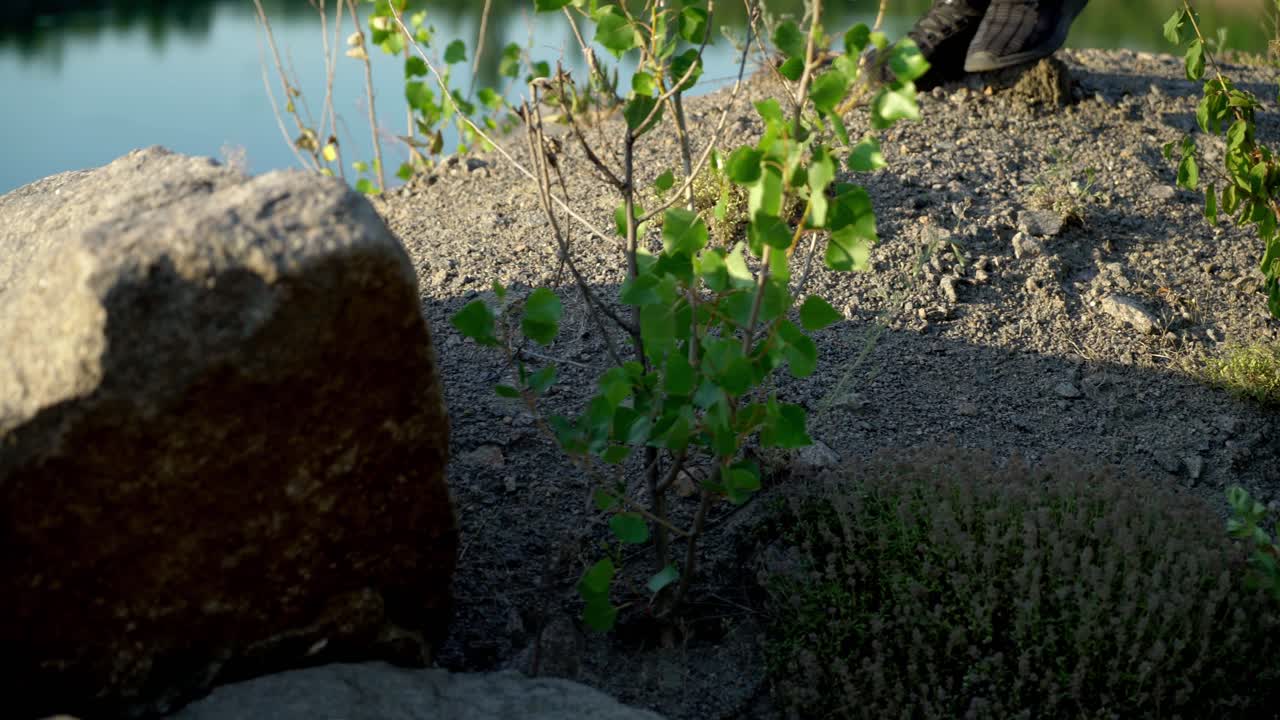 A man in black sneakers walks on stony ground along the lake on a sunny day of summer. Close-up.