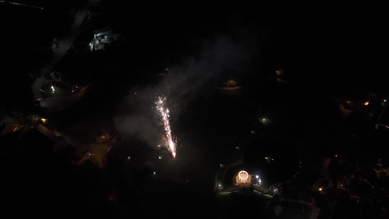 Fireworks bursting in the night sky over a lit pavilion and surrounding dark landscape.