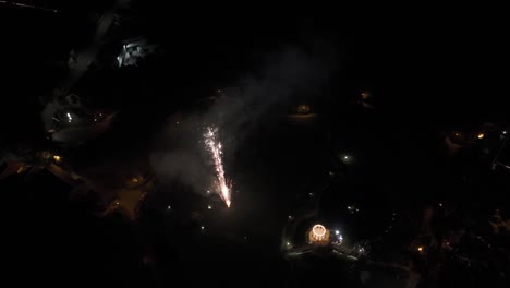 fireworks bursting in the night sky over a lit pavilion and surrounding dark landscape.