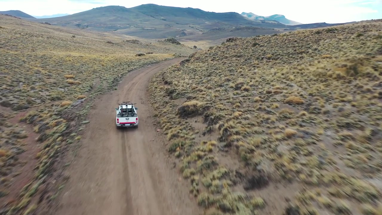 Ripresa con drone dall'alto di un veicolo 4x4 bianco che percorre un sentiero sterrato tortuoso attraverso una vasta valle di montagna a Bariloche, Argentina. Evidenzia viaggi remoti, esplorazione avventurosa e natura selvaggia.