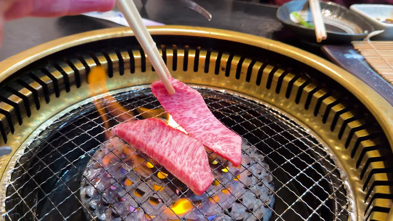 Bacchette posizionano manzo crudo marmorizzato su una griglia a carbone ardente in un ristorante giapponese di yakiniku.