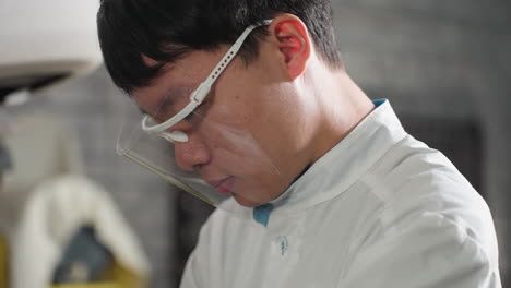 close-up of lab scientist wearing protective glasses with head bowed, in white lab coat, background featuring a mechanical workshop