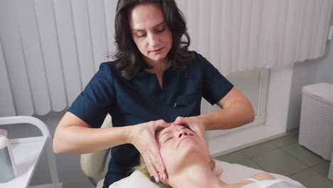 facialist massaging face of client lying peacefully on massage bed, client momentarily tenses then relaxes under skilled fingertips, spa environment with trolley, skincare bottles and ring light