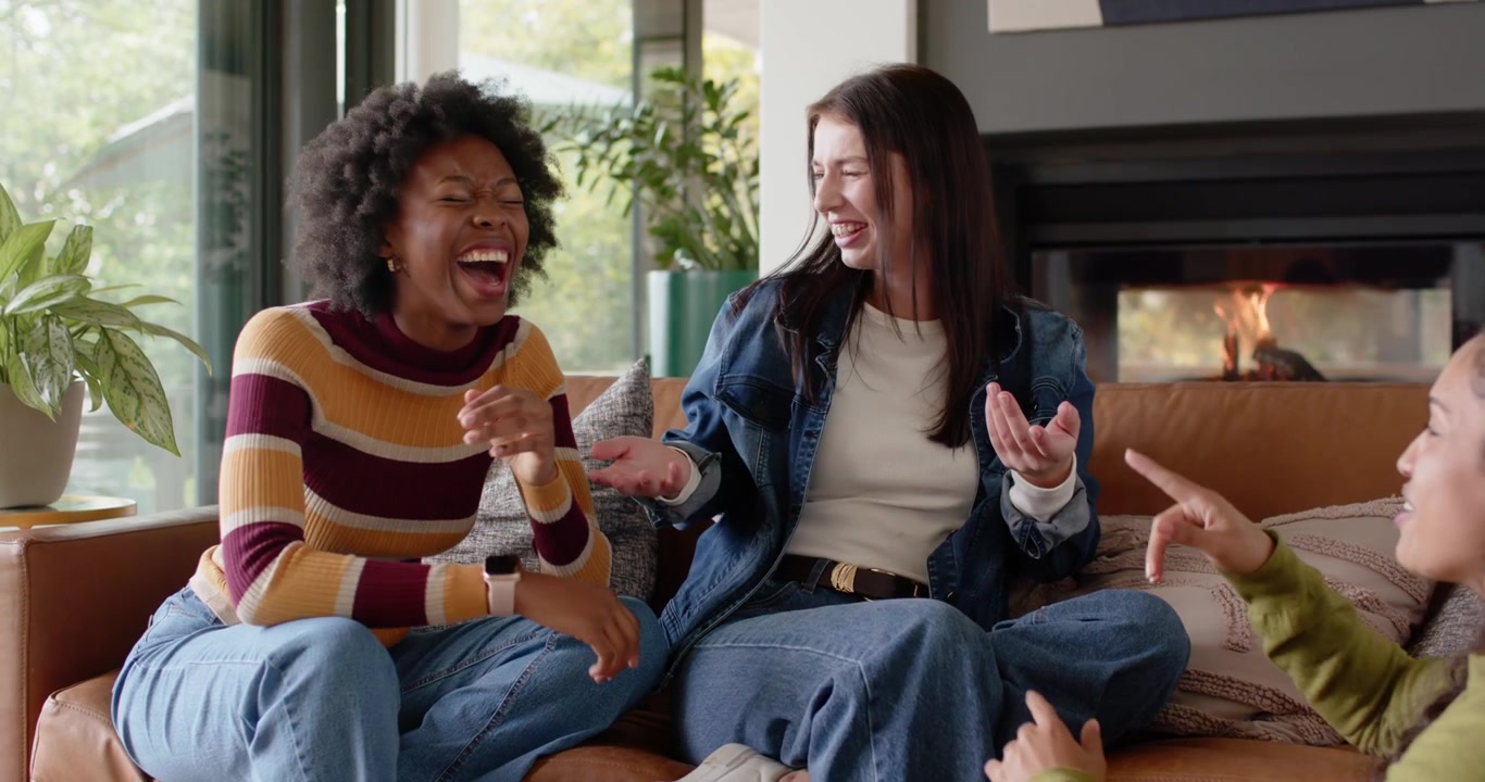 Diverse female friends mimicking left woman's gestures, laughing during lighthearted chat on sofa
