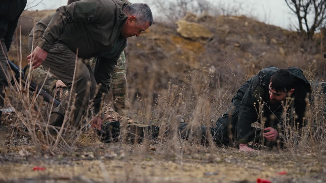 Four confident male experienced soldiers carry a wounded soldier in a camouflage uniform and a brunette man in dark green clothes stumbles dropping a wounded soldier during fighting in the steppe