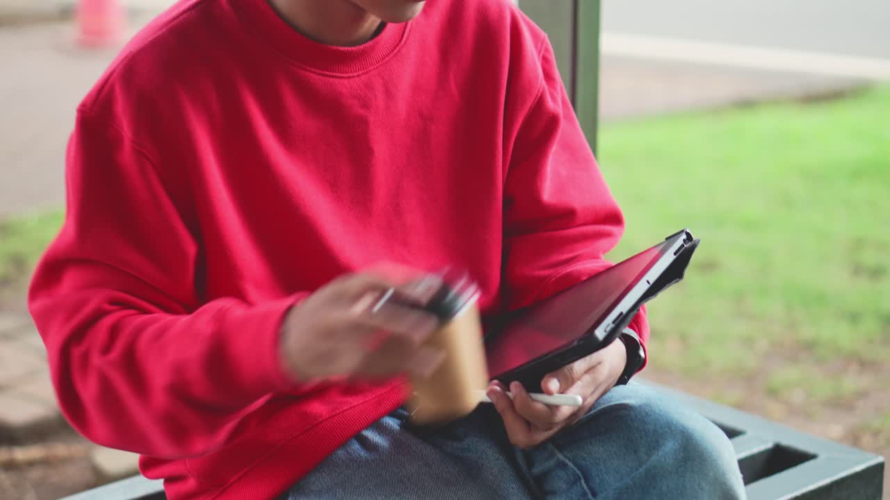 joven freelance masculino asiático usando auriculares y bebiendo café mientras trabaja en una computadora portátil en el parque