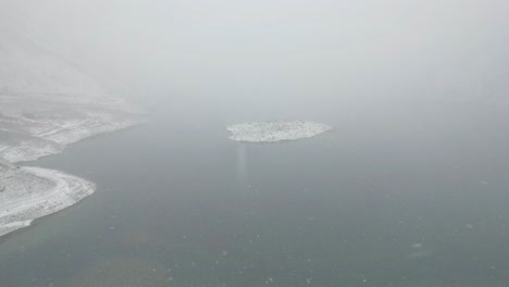 Aerial-View-Looking-Through-Dense-Misty-Foggy-Air-As-It-Snows-Over-Lake-In-Skardu