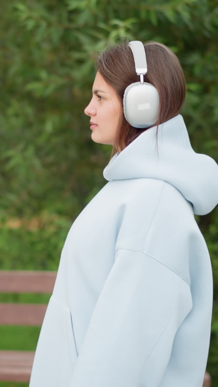 Bella donna in abbigliamento casual che passeggia in giardino indossando cuffie bianche, con uno sfondo che presenta fiori e una panchina di cemento, ideale per passeggiate all'aperto, natura o scene di relax.