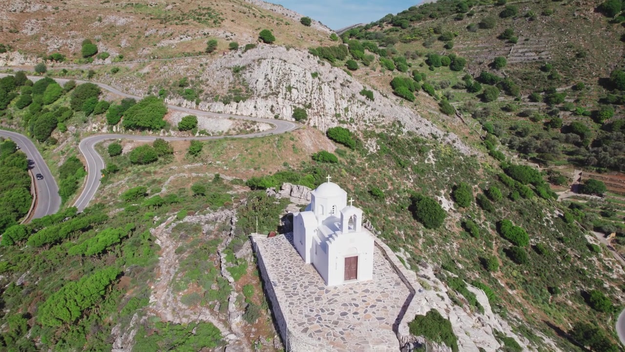 Cappella tradizionale greco-ortodossa arroccata sulla cima di Naxos. Campanile e cupola cicladici imbiancati a calce in mezzo alla serena eredità spirituale dell'Egeo, ripresa da drone aereo 4k.