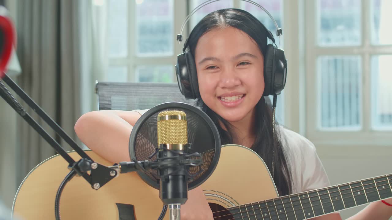 Over Shoulder View, Asian Kid Girl In Headphones Playing The Guitar And Singing Into Microphone While Recording Podcast With Boy Host In Studio