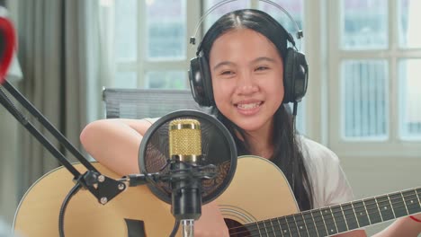 over shoulder view, asian kid girl in headphones playing the guitar and singing into microphone while recording podcast with boy host in studio