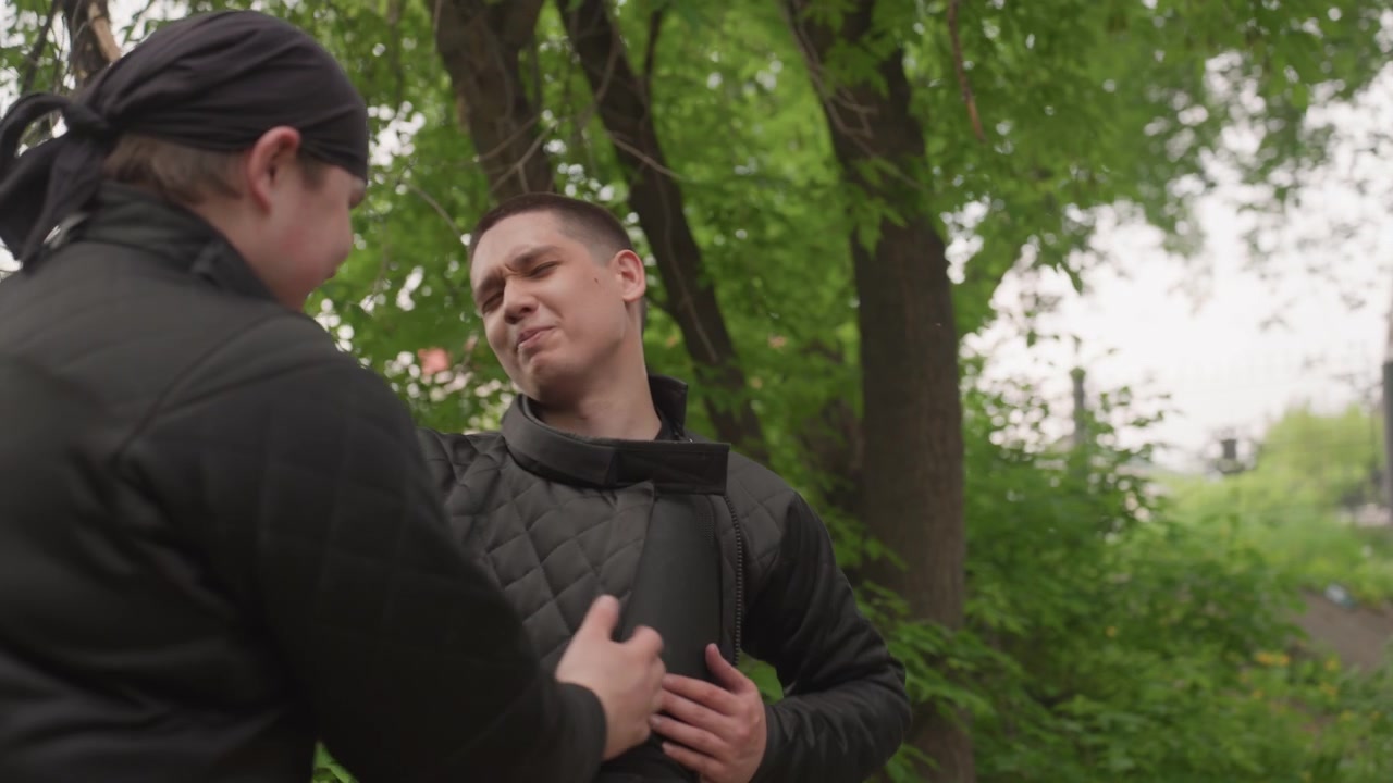 Close up of two men in black jackets outdoors one wearing padded vest. man with pad interacts closely as another men approaches and assists him through various gestures. Dense green foliage