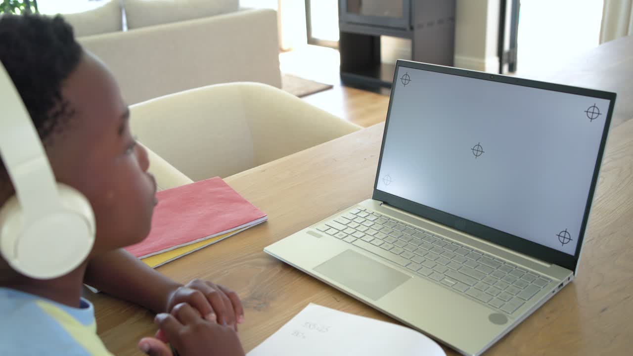 Áudio do laptop iniciando a reprodução, menino se inclinando para frente na mesa, anotando em aula on-line
