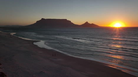 cape town south africa family people walking on sand windy surf wave table mountain drone aerial cinematic lions head sunset beach stunning mountain and ocean landscape orange gold sun backward motion