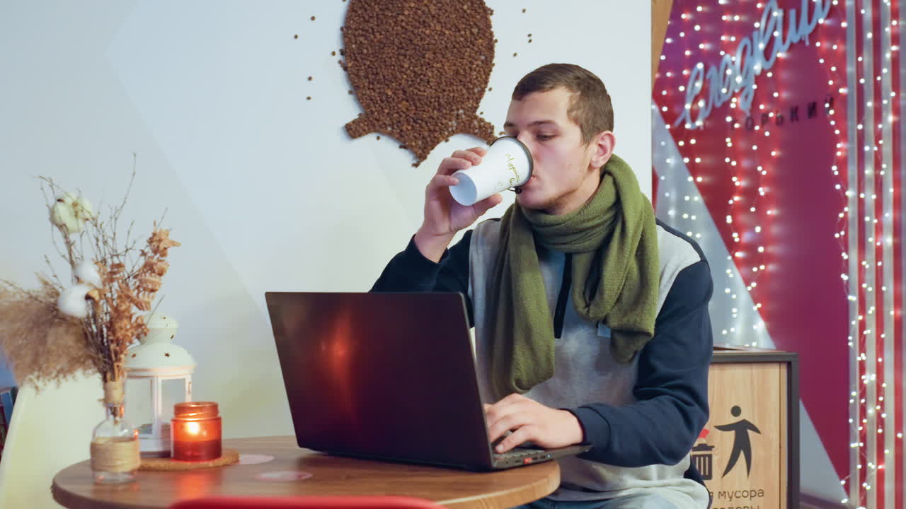 Uomo caucasico che beve caffè mentre lavora al laptop in un caffè decorato, la routine diurna mostra sorseggiare dalla tazza, controllare i messaggi, digitare e pianificare, candela e fiori secchi sul tavolo, concentrato.