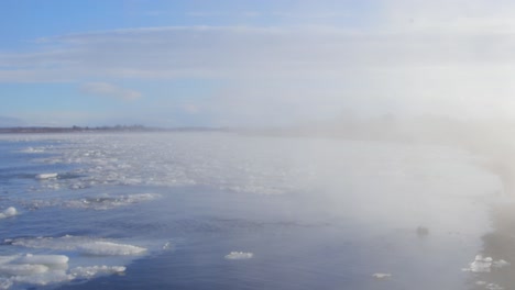 white smoke passes through the iceland blue lake with floating melting glaciers under brilliant blue sky - pan left shot