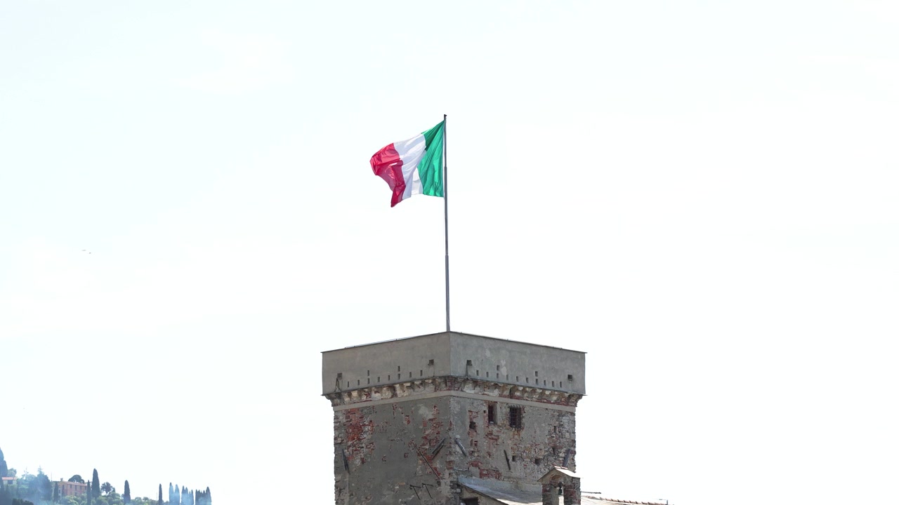 La bandiera italiana sventola in cima a un'antica torre di pietra sotto la piena luce del sole, mettendo in risalto i mattoni consumati e l'architettura medievale in un contesto di cielo sereno e tranquillo. Nessuna persona visibile.