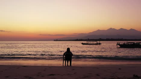 silhouette of girl sitting on chair over sandy beach, watching colorful sky after sunset with mountains horizon and anchored boats on calm lagoon, bali