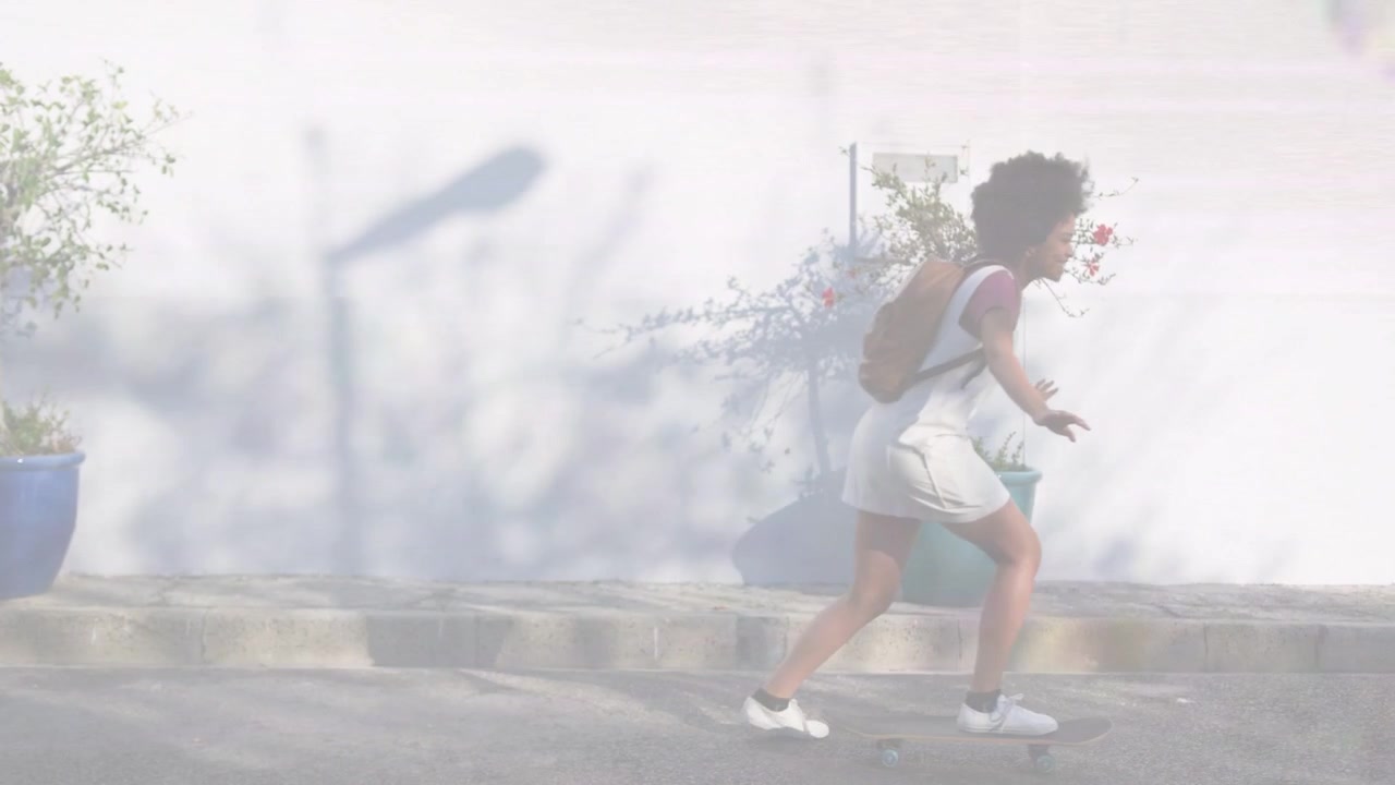 Wanita mendorong skateboard dan melewati pot tanaman biru di dekat dinding putih, melambat di dekat trotoar, dalam perjalanan.