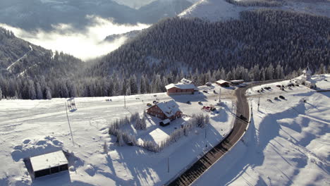 Straße-Vom-Gipfel-Der-Schneebedeckten-Berge,-Die-Durch-Einen-Ort-Mit-Ein-Paar-Häusern-Und-Einer-Kirche-Führt,-Luftaufnahme-Aus-Rumänien,-Rodna-Gebirge