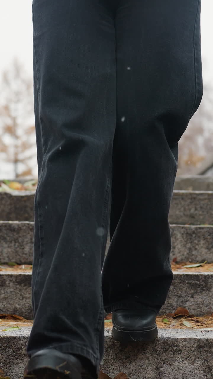 Legs of person walking down stone steps during light snowfall, wearing black jeans and black boots, with colorful autumn leaves scattered along the path