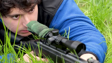 male lying on the grass, aiming through his weapon sight