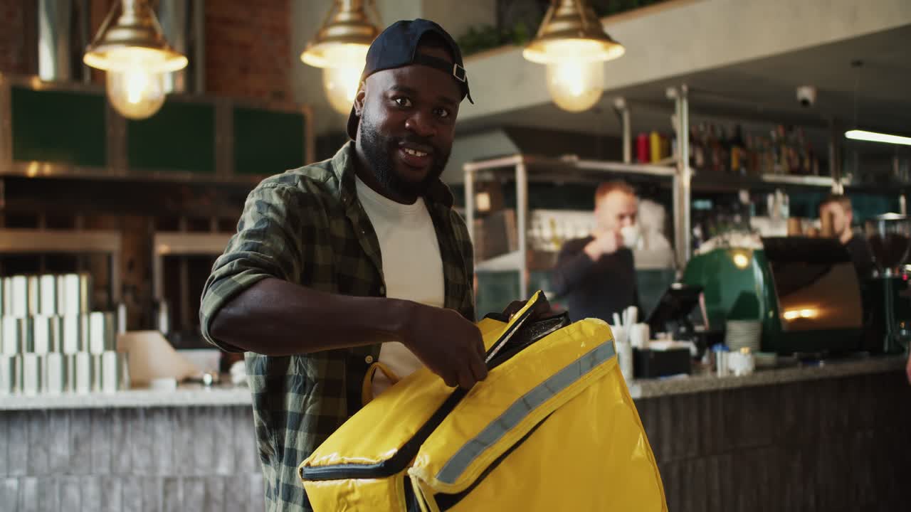 A food delivery man in a plaid shirt and black cap poses and smiles at a doner market. A man opens a special thermal bag for delivery