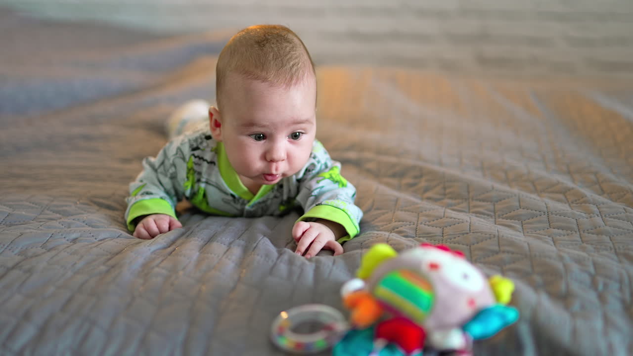 Tiny boy lies on the bed and toy is in front of him. Cute kid moving arms and legs actively, making funny faces. Grey backdrop in blur.