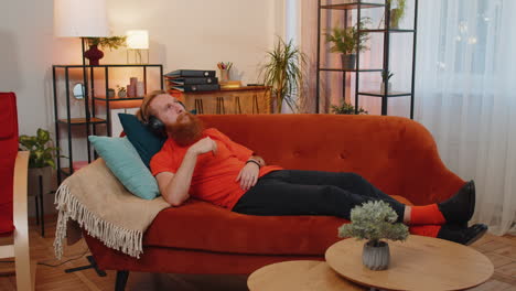 happy young man in wireless headphones relaxing lying on sofa at home listening favorite music