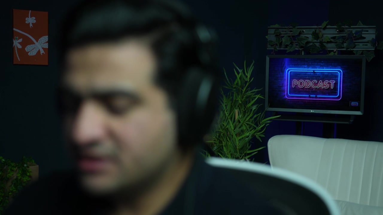 Blurred foreground of male host putting on headphones in a podcast studio with neon sign on screen behind. Audio broadcasting workflow, sound monitoring and media recording concept