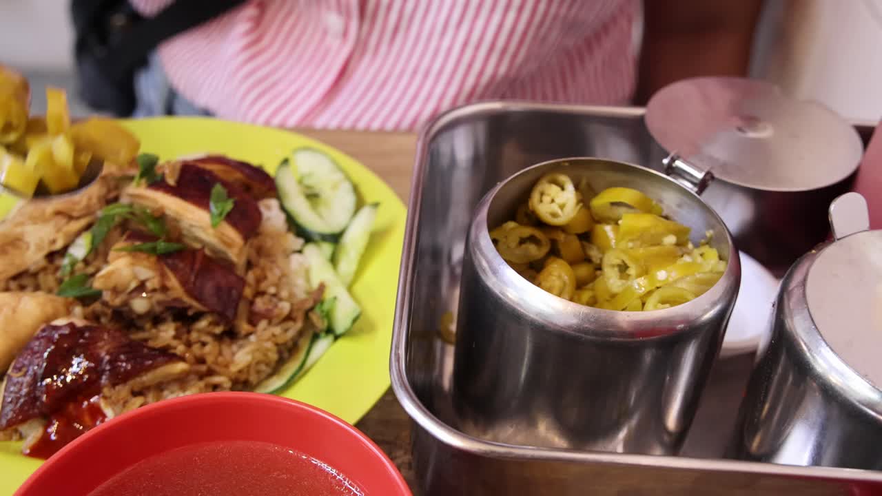 A person spoons pickled chili from a metal container onto a plate of roasted rice and meat, under bright indoor lighting with a close-up perspective