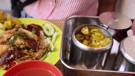 a person spoons pickled chili from a metal container onto a plate of roasted rice and meat, under bright indoor lighting with a close-up perspective