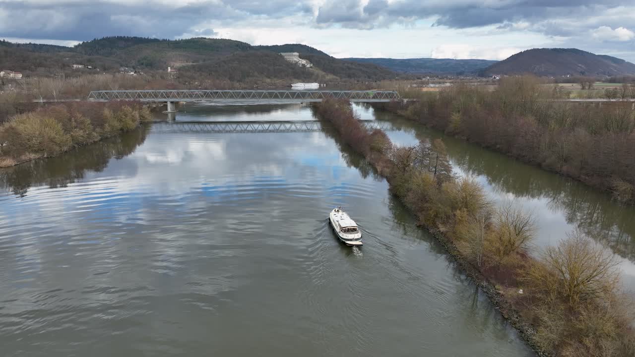 A beautiful aerial shot of a passenger boat cruising on a wide river, heading towards a classic metal truss bridge in the German countryside
