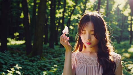긴 드레스를 입은 젊은 여성이 조용한 숲에서 손가락에  나비를 들고있는 나무를 통해 태양빛이 필터링됩니다.