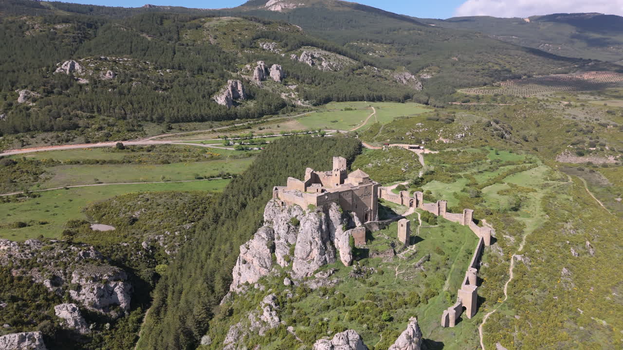 Slow drone orbit around an ancient stone castle atop a rugged cliff, surrounded by dense forest, green fields and distant mountain ranges under a clear blue sky