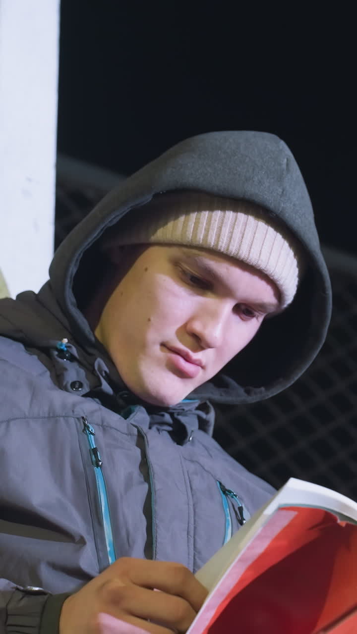 Young man in hoodie and beanie leaning against metallic goalpost, focused reading book outdoors at night with mesh fence background